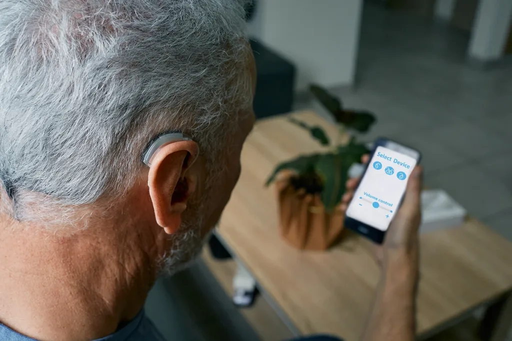 An older man wearing a Bluetooth hearing aid looking down at his smartphone pairing them together via a wireless Bluetooth connection