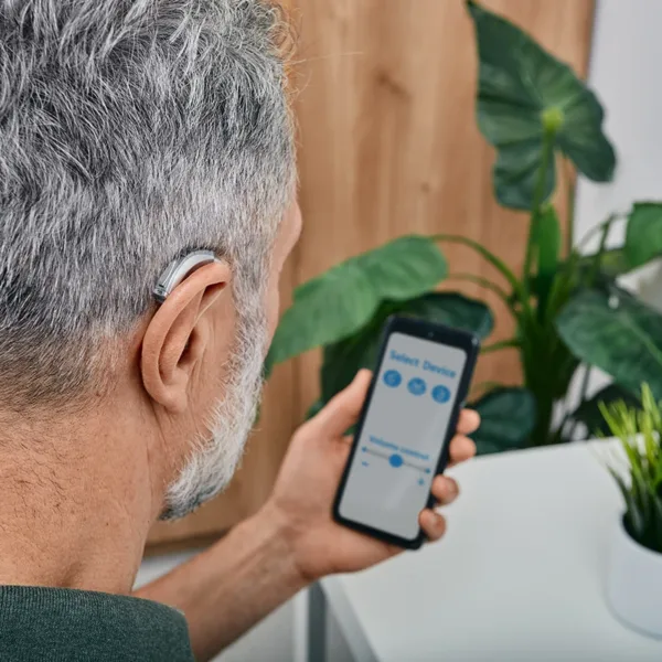 A man with grey hair, wearing a BTE style AI hearing aid, looking down at him smartphone at an AI hearing aid app with virtual assistant features