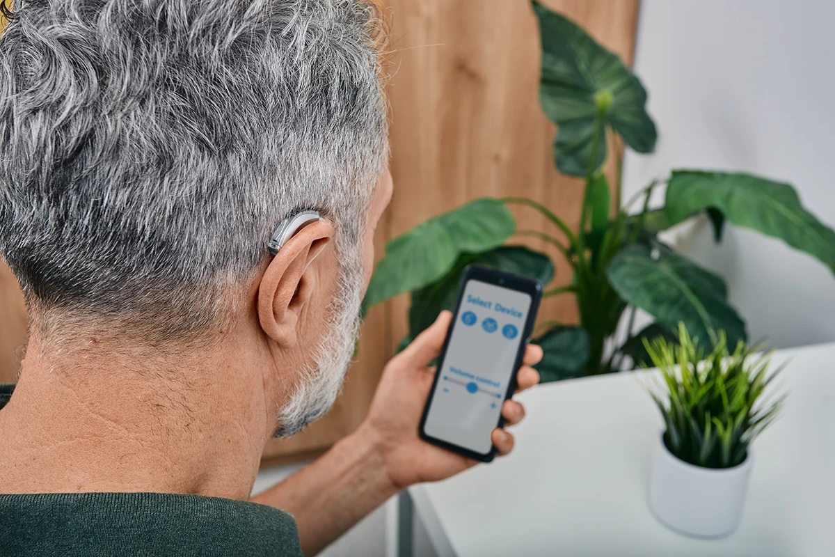 A man with grey hair, wearing a BTE style AI hearing aid, looking down at him smartphone at an AI hearing aid app with virtual assistant features