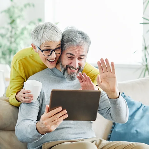 An older, retirement age couple sitting in their living room holding an iPad tablet up, Facetiming with a loved one, demonstrating how to use Siri with hearing aids