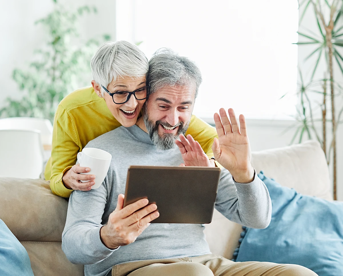 An older, retirement age couple sitting in their living room holding an iPad tablet up, Facetiming with a loved one, demonstrating how to use Siri with hearing aids