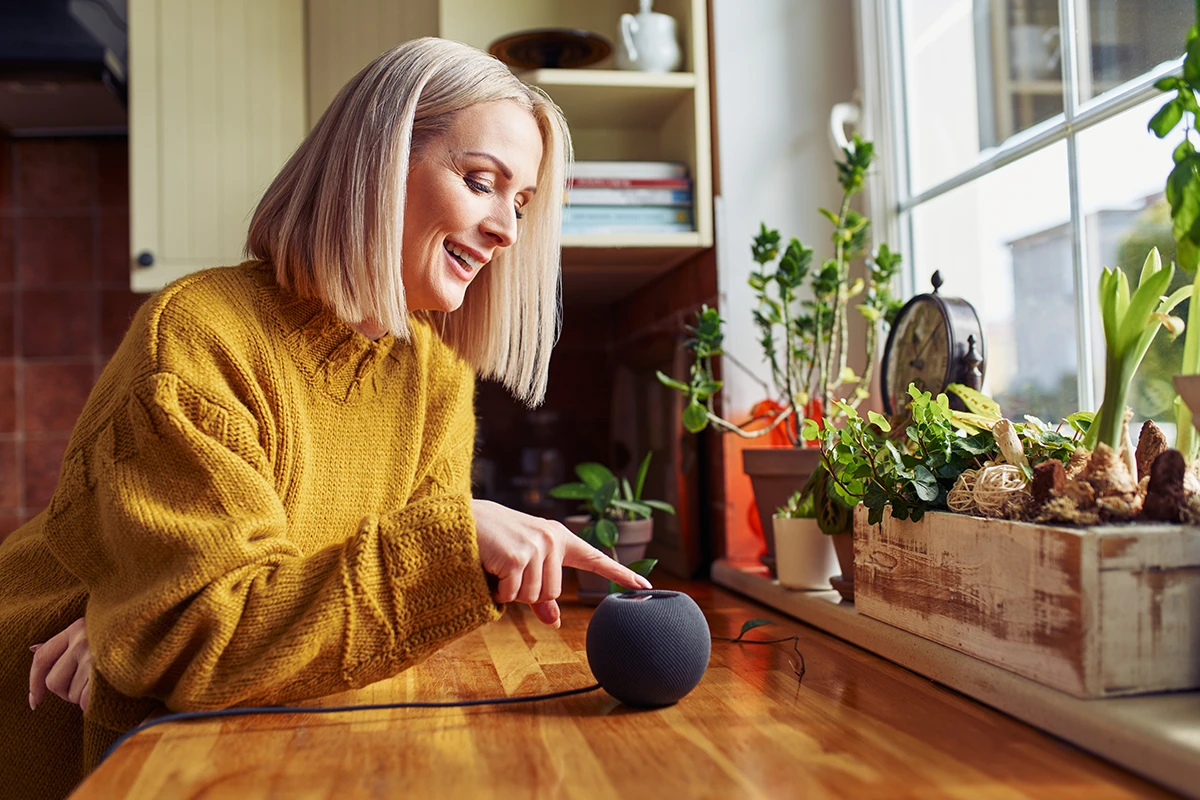 Happy mature woman connecting Alexa to hearing aids via a Bluetooth smart speaker at home in a kitchen