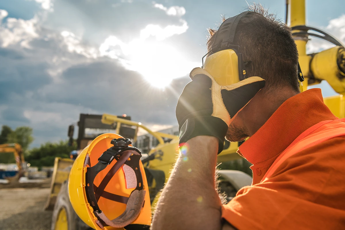 A person outdoors at a construction site, standing near heavy machinery, wearing double hearing protection to prevent noise-induced hearing loss at work