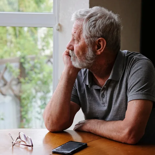 A mature, senior man, who appears to be sad, lonely, or depressed, looking in distance out of window, symbolizing age-related hearing loss and social isolation in older adults