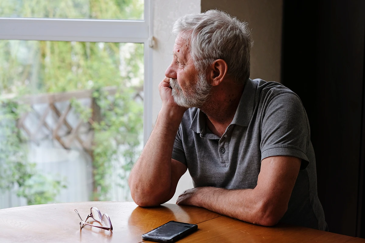 A mature, senior man, who appears to be sad, lonely, or depressed, looking in distance out of window, symbolizing age-related hearing loss and social isolation in older adults