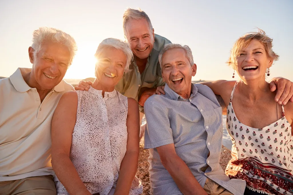 A friend group of five older adults seniors sitting on a beautiful beach at sunset, happily laughing and talking with each other, symbolizing how hearing aids combat social isolation in older adults.