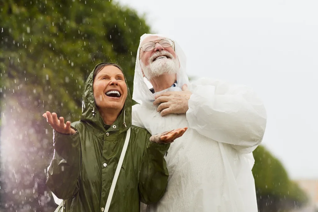 A senior man and women wearing rain jackets happily standing in the rain together enjoying the rain drops as they fall on the faces and hands, symbolizing the peace of mind that comes with wearing waterproof hearing aids.