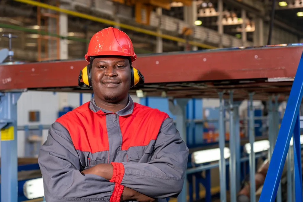 A young, male, industrial worker in red helmet, coveralls, and hearing protection equipment to prevent noise-induced hearing loss at work in a factory