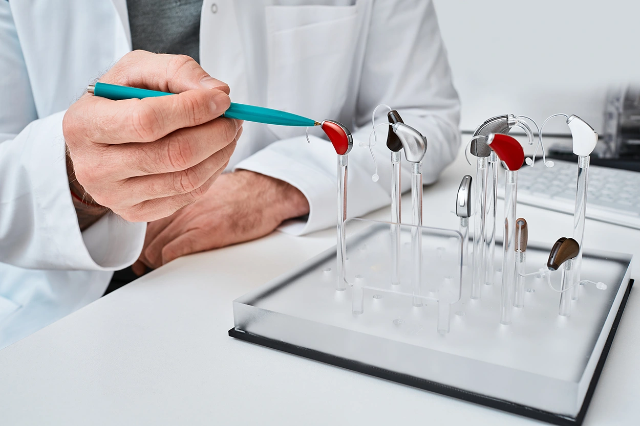 A hearing health care specialist pointing to a hearing aid displaying multiple different styles and types of hearing aids, explaining how long hearing aids last