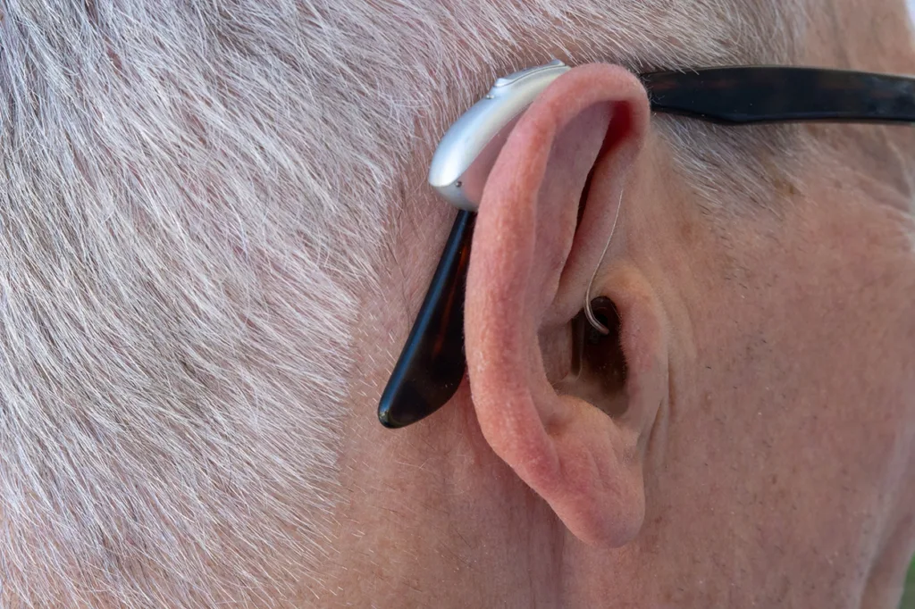 Close-up of the ear of a grey-haired man wearing eyeglasses and a modern compact BTE prescription hearing aid.