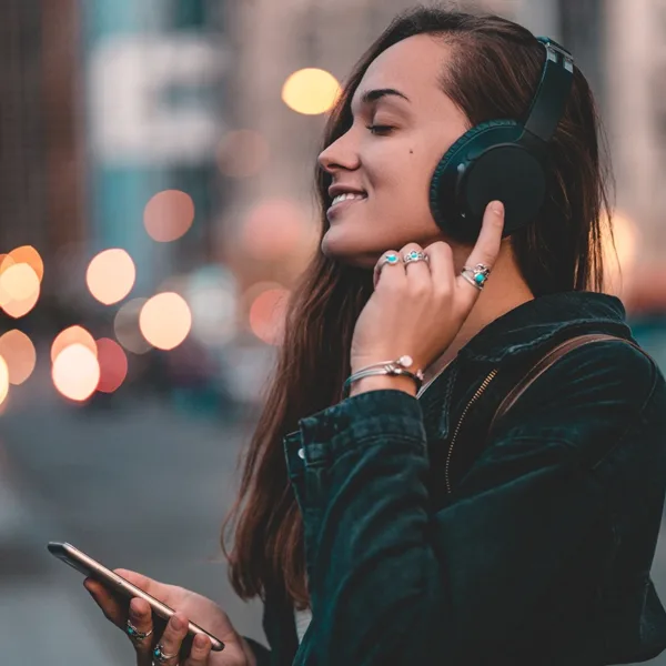 Young, happy, stylish, woman listening to music on a black wireless headphone while walking around the city.