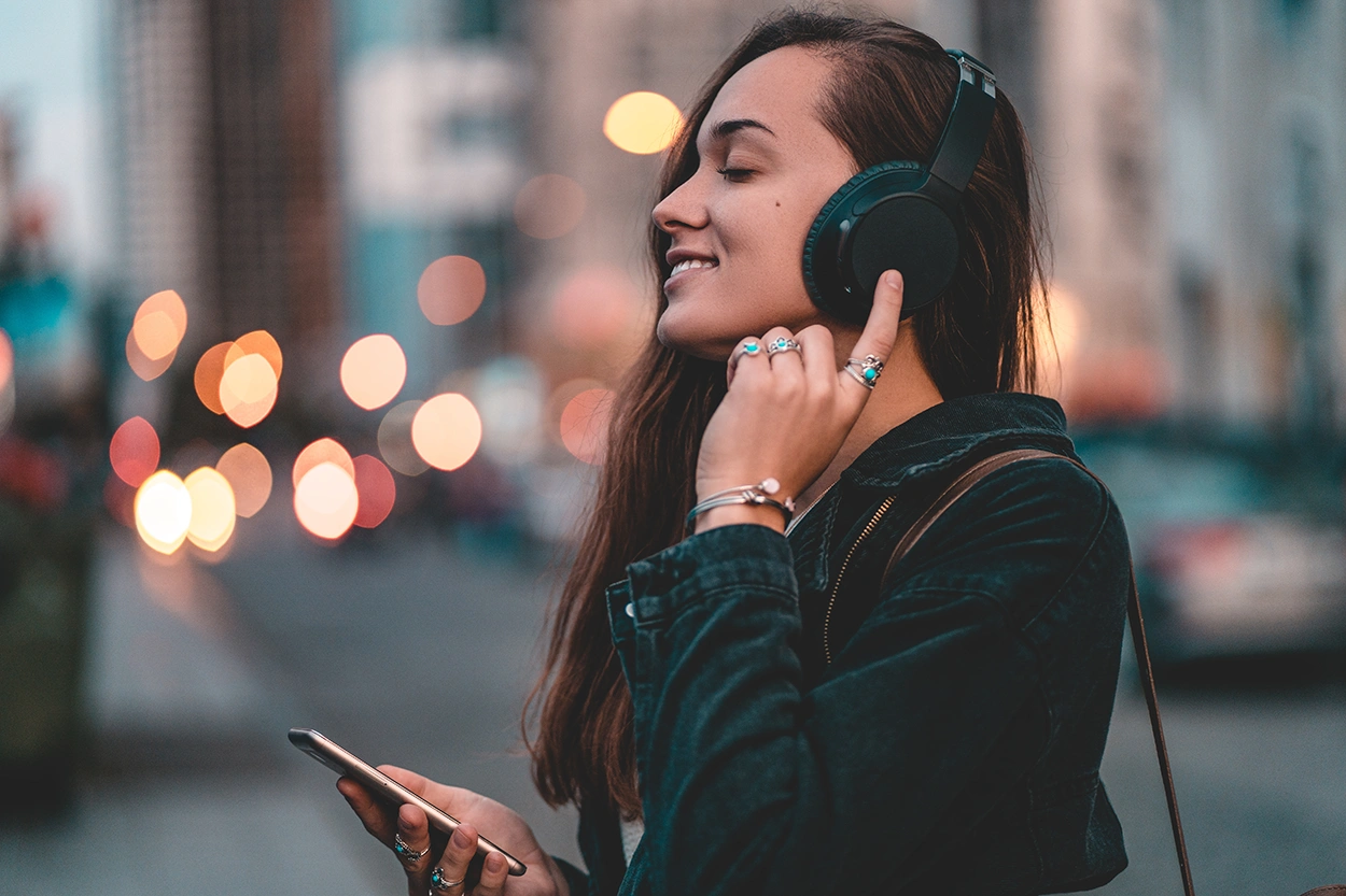 Young, happy, stylish, woman listening to music on a black wireless headphone while walking around the city.