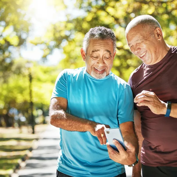 2 older adult men with short grey hair wearing workout clothes, smiling and looking at a fitness tracking hearing aids app on a smartphone