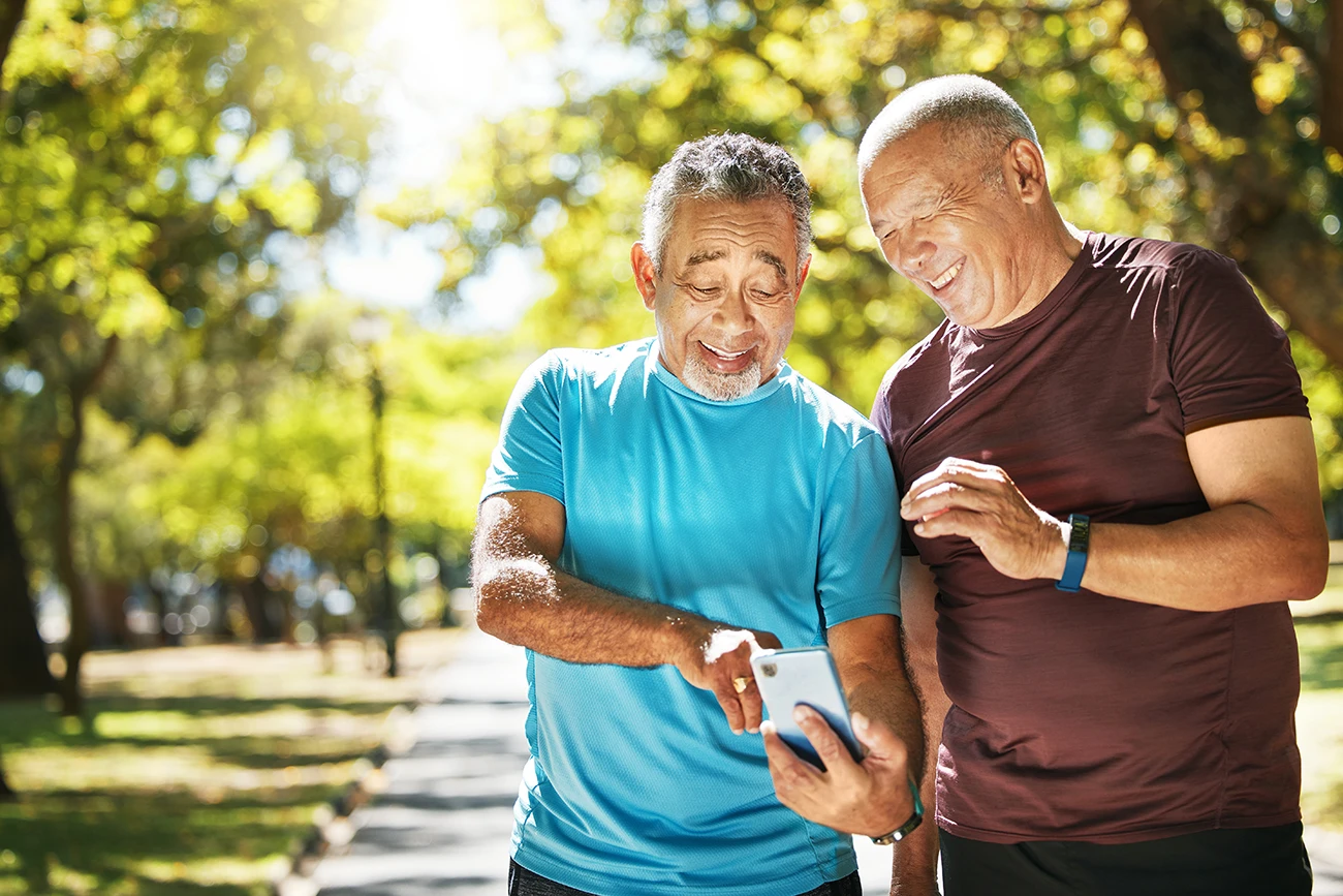 2 older adult men with short grey hair wearing workout clothes, smiling and looking at a fitness tracking hearing aids app on a smartphone