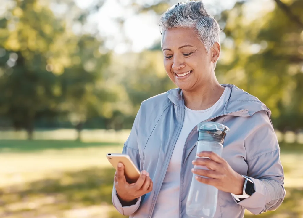 an older adult women with short grey hair wearing workout clothes, smiling and looking at her fitness tracking hearing aids app on her smartphone