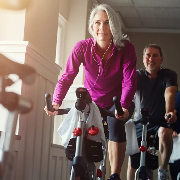 Adult woman with grey hair sitting on an exercise bike during a spin class, wearing earbuds, protecting hearing at the gym